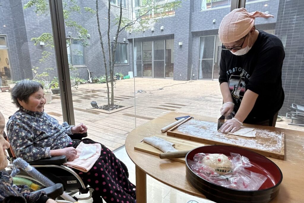 入居者と職員が一緒に調理や食事準備をしている様子。窓を通じて庭が見える。